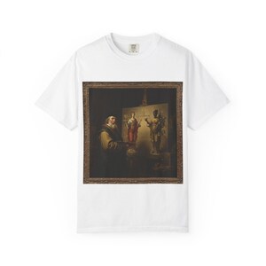 May include: White t-shirt featuring a framed painting of a man painting a canvas with two figures. The painting has a dark brown frame and depicts a man with a beard and a canvas with two figures. The t-shirt has a crew neck.