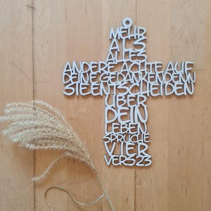 May include: A white wooden cross with German text, including the words "Mehr als alles" and "Vier Vers 23". A dried plant stem with feathery seed heads is to the left of the cross. The background is a light brown wooden surface.