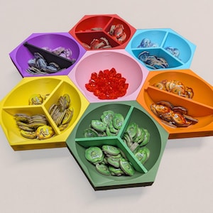 Puede incluir: Un organizador de juegos hexagonal multicolor con siete compartimentos. Cada compartimento es de un color diferente: morado, rojo, azul, naranja, amarillo, rosa y verde. Los compartimentos contienen piezas de juego de varias formas y colores.