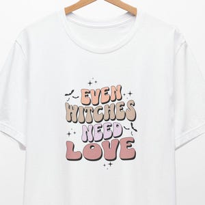 Op de afbeelding: Wit t-shirt met de tekst "EVEN WITCHES NEED LOVE" in een retrostijl. De tekst is in roze, perzik en bruin, met sterren. Het t-shirt hangt aan een houten hanger.