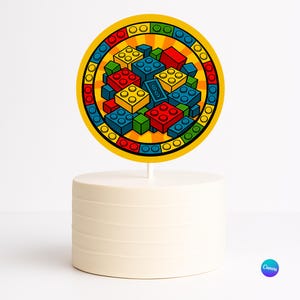 Puede incluir: Un adorno redondo para pastel con una colorida ilustración de bloques de construcción de juguete en rojo, azul, amarillo y verde. El adorno está sobre un palito blanco, colocado encima de un pastel blanco con estrías horizontales.