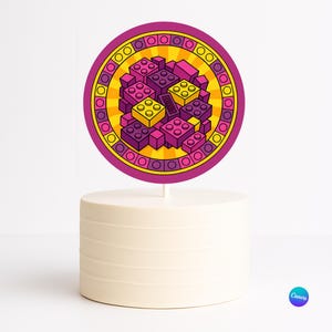 Puede incluir: Un adorno redondo para pastel con un diseño colorido de bloques de construcción de juguete en tonos morado, rosa y amarillo. El adorno está sobre un pastel blanco con estrías horizontales.