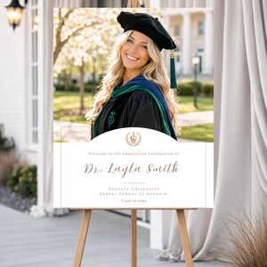 Puede incluir: Un letrero de celebración de graduación sobre un caballete. El letrero presenta una foto de una graduada sonriente con birrete y toga. El texto en el letrero dice "Bienvenida a la Celebración de Graduación de la Dra. Layla Smith" e incluye el nombre de la universidad y el año.