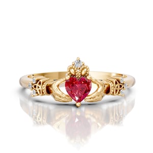 Könnte beinhalten: Ein goldfarbener Claddagh-Ring mit einem herzförmigen roten Edelstein. Der Ring zeigt Hände, die das Herz halten, eine Krone darüber und kleine Akzentsteine. Der Ring ist vor einem weißen Hintergrund platziert.