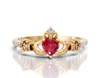 May include: A gold-colored Claddagh ring with a heart-shaped red gemstone. The ring features hands holding the heart, a crown above, and small accent stones. The ring is set against a white background.