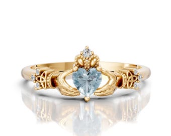 May include: A gold-colored Claddagh ring with a heart-shaped, light blue gemstone. The ring features hands holding the heart, a crown above, and Celtic knot details on the sides. Small clear stones are set in the crown and on the sides.