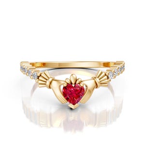 May include: A gold-colored Claddagh ring with a heart-shaped red gemstone at the center. The ring features two hands holding the heart, with a crown above. Small clear gemstones are set along the band.