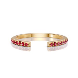 May include: A gold-colored open ring with a row of small, bright red gemstones set along the outer band. The ring has a gap in the center, creating a unique design. The gemstones are evenly spaced and add a pop of color.