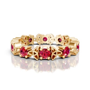 May include: A gold ring with a repeating pattern of red gemstones and Celtic knot designs. The ring's design incorporates round gemstones and intricate knotwork, creating a blend of elegance and cultural symbolism. The ring is displayed on a white background.