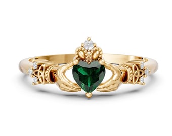 Pode incluir: Um anel Claddagh de ouro com uma pedra preciosa verde-escura em forma de coração no centro, segurada por duas mãos esculpidas. A banda é adornada com detalhes metálicos intrincados, pequenas pedras claras e um motivo de coroa.