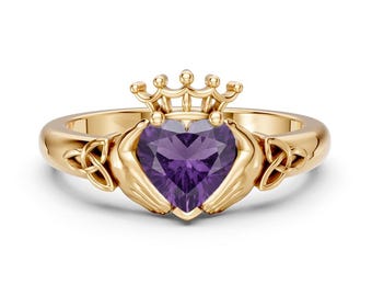 May include: A gold Claddagh ring featuring a central heart-shaped purple amethyst gemstone. The design includes two hands holding the heart, a crown resting on top, and intricate Celtic knotwork details on both sides of the polished gold band.