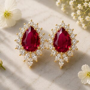 May include: A pair of gold-toned stud earrings featuring a large, pear-shaped deep red gemstone at the centre, surrounded by a halo of small, clear, round and marquise-cut white crystals arranged in a floral-inspired, pointed cluster design.