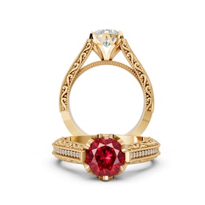 May include: A gold ring featuring a round, deep red gemstone set in a prong setting. The band is decorated with intricate scrollwork and small, clear accent stones along the sides. The ring is shown from both a front and a side perspective.