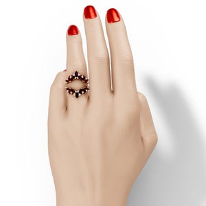 May include: A gold-toned ring with a double-band design, adorned with red and white gemstones. The ring is set with a series of small, round, and marquise-cut stones. The ring is on a hand with red nail polish.