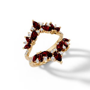 May include: A gold ring featuring a cluster of marquise-cut, deep red gemstones and small, round, white gemstones. The ring's design is a stylized floral or leaf pattern, with the gemstones set in a symmetrical arrangement.