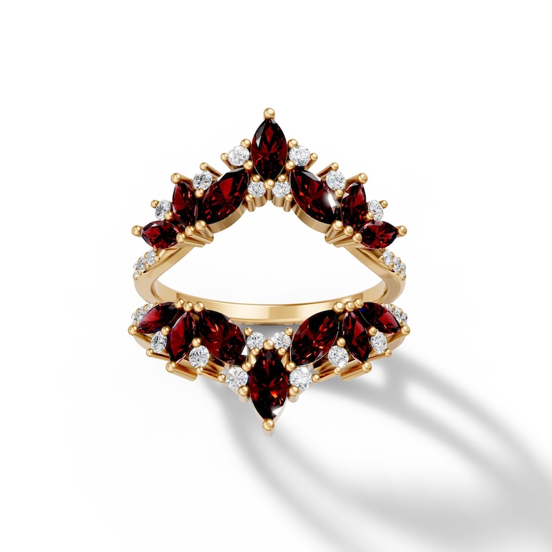 May include: A gold ring with a unique design. The ring features a series of marquise-cut, deep red gemstones and small, round, white gemstones. The gemstones are set in a floral or leaf-like pattern. The ring is set against a white background.