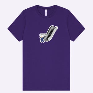 May include: A purple t-shirt featuring a cartoon graphic of an escalator with a person in a suit and the word "UPPIES!!!" in a speech bubble. The shirt is made of a soft material.