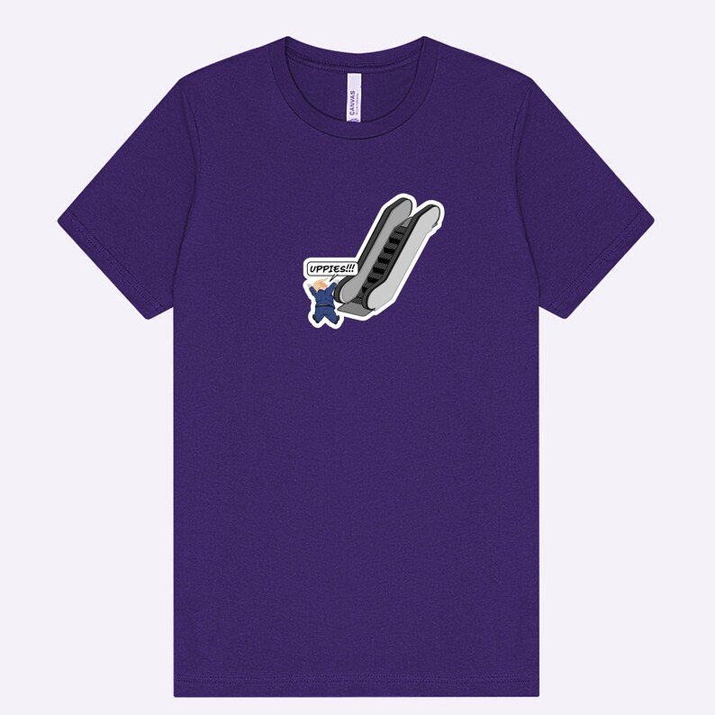 May include: A purple t-shirt featuring a cartoon graphic of an escalator with a person in a suit and the word "UPPIES!!!" in a speech bubble. The shirt is made of a soft material.