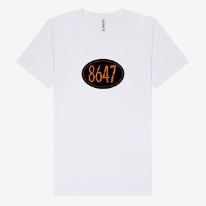 May include: White V-neck t-shirt with a black oval design in the center. The oval contains the number "8647" in a fiery orange font. The shirt has short sleeves and a relaxed fit.