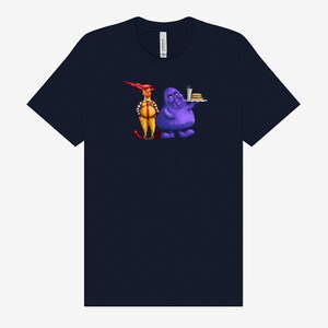 May include: Navy blue t-shirt featuring a cartoon illustration of two characters. One character is dressed in a yellow and red outfit, and the other is a purple blob holding a tray with a burger and drink.