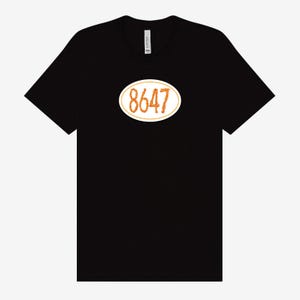 May include: Black t-shirt featuring an oval design with the number "8647" in a textured orange font, outlined in white. The shirt is made of a soft material and has a crew neck.