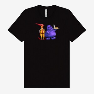 May include: Black t-shirt featuring a cartoon illustration of two characters. One character is dressed in a yellow and red uniform, and the other is a purple blob holding a tray with a burger and drink.