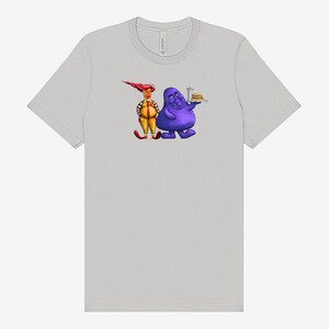 May include: Light gray t-shirt featuring a cartoon illustration of two characters. One character is purple and holding a tray with a burger. The other character is yellow and red. The shirt is made of a soft material.