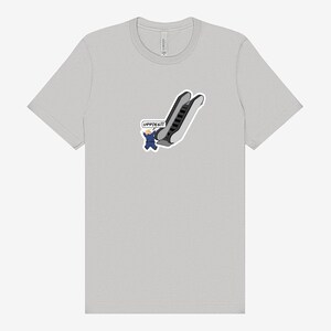 May include: A light gray t-shirt featuring a cartoon of a person in a blue suit falling down an escalator. The text "UPPIES!!!" is in a speech bubble. The shirt is a basic crew neck.