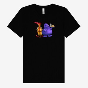 May include: Black V-neck t-shirt featuring a cartoon illustration of two characters. One character is dressed in a yellow and red outfit, and the other is a purple blob holding a tray with a burger and drink.