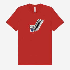 May include: Red t-shirt featuring a cartoon graphic of a person in a blue suit falling down an escalator. The text "UPPIES!!!" is in a speech bubble. The shirt is made of a soft material.