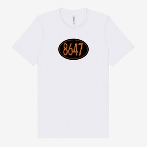 May include: White t-shirt with a black oval design in the center. The oval contains the number "8647" in a stylized orange font. The shirt has a crew neck and short sleeves.