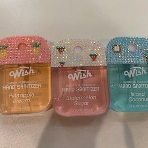 May include: Three hand sanitizer bottles with bedazzled tops. The bottles are labeled Pineapple Dream, Watermelon Sugar, and Island Coconut. Each bottle has a different color of liquid and decorative embellishments.