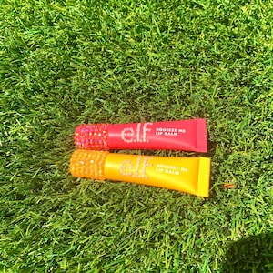 May include: Two tubes of e.l.f. Squeeze Me Lip Balm rest on a bed of green grass. One tube is red with a bedazzled pink cap, and the other is yellow with a bedazzled orange cap. The text "e.l.f." and "Squeeze Me Lip Balm" are printed on the tubes.