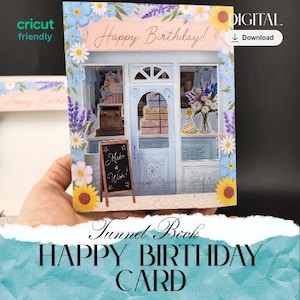 May include: A 3D pop-up Happy Birthday card with a shop front design. The card features a blue and white facade, floral accents, and the words "Happy Birthday!" and "Make a Wish!". The card is a tunnel book design and is labeled "Happy Birthday Card".