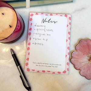 May include: A rectangular notepad with a pink border featuring a repeating strawberry pattern. The page is headed with the word Notes and contains a handwritten checklist. At the bottom, the text reads: Charm is deceitful and beauty is vain but a woman who fears the Lord is to be praised Prov 31:30.