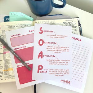 May include: A white card with the acronym SOAP printed in large red letters, explaining the Bible study method: Scripture, Observation, Application, and Prayer. The card rests on an open Bible with handwritten notes, a blue mug, and a pen.