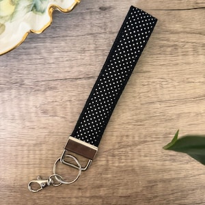 May include: Black and white polka dot wristlet keychain with silver-tone hardware. The wrist strap is made of a sturdy fabric and features a repeating pattern of small white dots. The keychain includes a split ring and a lobster clasp for easy attachment.