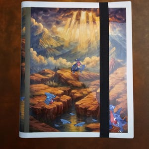 Puede incluir: Un archivador de tarjetas con borde blanco que presenta una ilustración de paisaje con un río, montañas y un cielo nublado con rayos de sol. Varios personajes de dibujos animados azules y rojos son visibles. Una banda elástica negra asegura el archivador.