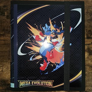 Może przedstawiać: Czarny segregator na karty z żywą ilustracją niebiesko-czerwonej postaci z żółto-pomarańczowymi wybuchami energii. Segregator posiada czarne zapięcie na gumkę, a u dołu widnieje napis "MEGA EVOLUTION".
