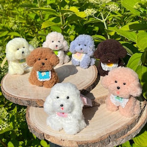 May include: A collection of plush toy poodle dogs in various colors, including white, brown, and blue. Each dog has a bib and is posed on a wooden slice. The background features green foliage.