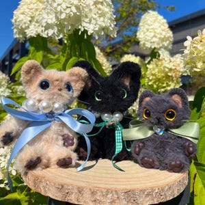 May include: Three plush cat figurines with large, expressive eyes and pearl necklaces. One is tan with a blue ribbon, another is black with a green ribbon, and the third is gray with a green ribbon. They sit on a wooden slice.