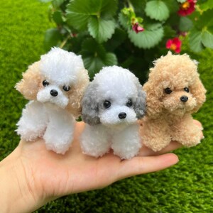 May include: Three small, fluffy toy poodles in shades of white, tan, and gray. The poodles have black eyes and noses, and are posed on a green background. The toys are made of a soft, textured material.