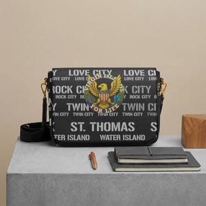 May include: A black messenger bag featuring a repeating white text pattern of LOVE CITY, ROCK CITY, TWIN CITY, ST. THOMAS, and WATER ISLAND. A circular emblem in the center displays a golden eagle with the text VIRGIN ISLANDS FOR LIFE.