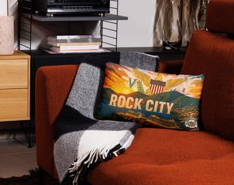 May include: A rectangular pillow with a Rock City design featuring a sunset, mountains, and a crest. The pillow is on a rust-colored couch with a black and white checkered throw blanket.