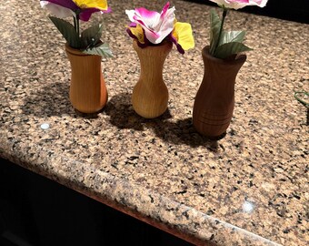 May include: Three small wooden vases, each holding a colorful artificial flower. The flowers have yellow, purple, and white petals. The vases are different shades of brown and sit on a speckled countertop.