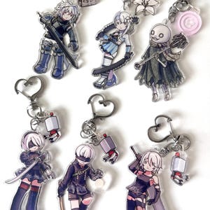 May include: A collection of six acrylic keychains featuring anime-style characters with heart-shaped clasps. Each keychain includes a small secondary charm, such as a book, a flower, a button, or a small robot, attached to the silver-toned metal ring.