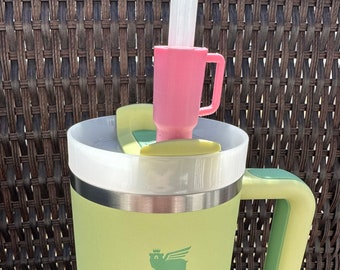 May include: A green Stanley travel mug with a black logo and a green handle. The mug has a pink lid with a straw.