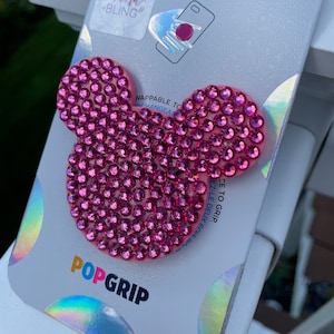 Puede incluir: Agarre para teléfono PopGrip rosa con un diseño de Mickey Mouse rosa brillante. El agarre está hecho por PopSockets y presenta el texto "PopGrip" y "adora BLING" en el empaque.