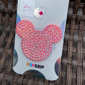 Puede incluir: Agarre para teléfono PopGrip rosa con un diseño de Mickey Mouse brillante e iridiscente. El empaque presenta el texto "POPSOCKETS" y "adora BLING".