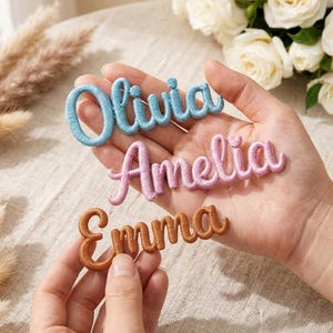 May include: Three embroidered name patches: "Olivia" in light blue, "Amelia" in pink, and "Emma" in brown. The names are in a cursive font, with a 3D effect. The patches are held in a person's hand, with a soft background.